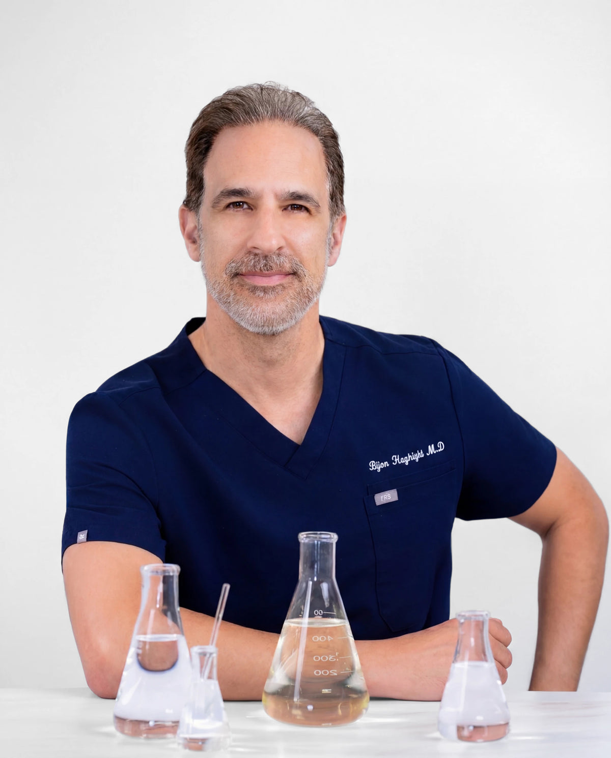 Dr. Bijan Haghighi, MD, in navy scrubs at a clean white surface with chemistry beakers.